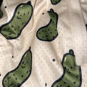 May include: Close-up of a fabric with a repeating pattern of green cartoon pickles with black outlines and smiling faces. The background is a light beige color with small black dots and a mesh-like texture.