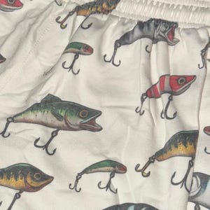 May include: White cotton fabric with a repeating pattern of fishing lures and fish. The lures are in various colors, including red, green, and yellow. The fish are all in shades of brown and green.