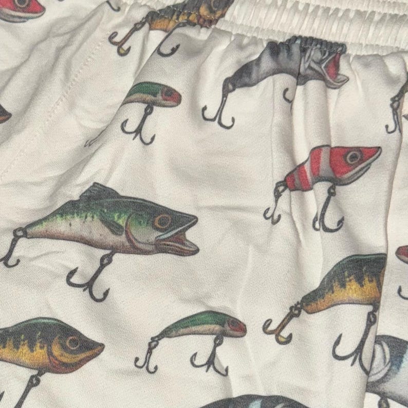 May include: White cotton fabric with a repeating pattern of fishing lures and fish. The lures are in various colors, including red, green, and yellow. The fish are all in shades of brown and green.