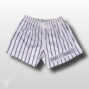 May include: White athletic shorts with vertical navy blue stripes. The shorts have a drawstring waist and the text "GOAT STRENGTH" in a navy blue rectangular box on the lower right leg. The shorts are made of a mesh material.