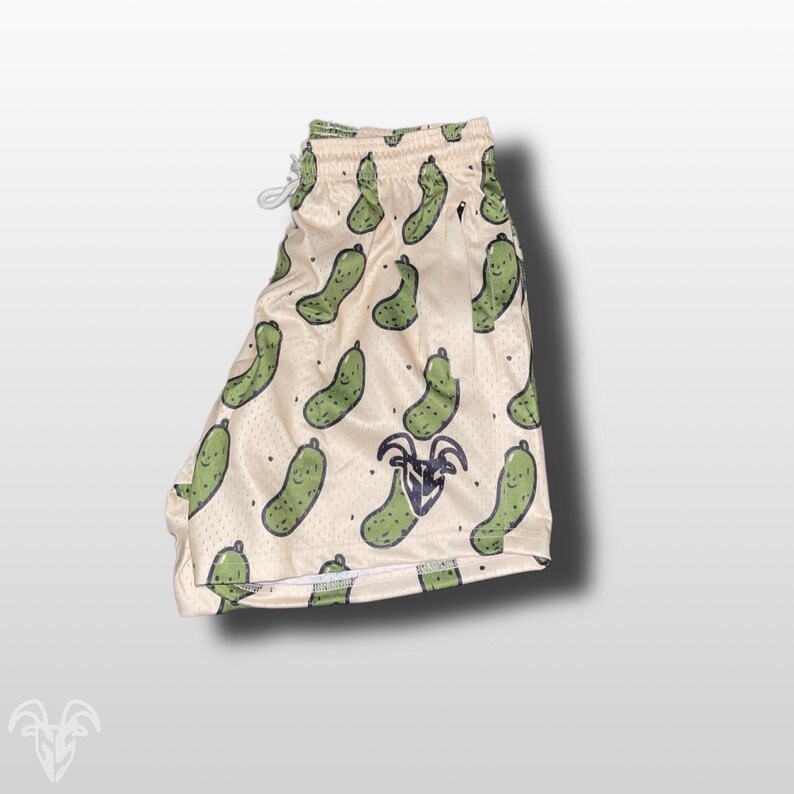 May include: Cream-colored mesh shorts with a repeating pattern of green cartoon pickles. The shorts have a drawstring waistband and a logo near the bottom. The shorts are designed for casual wear.