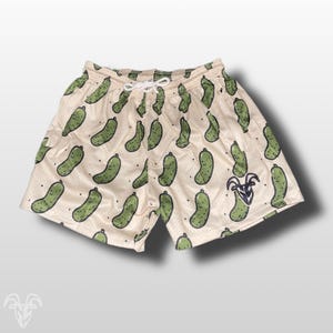 May include: Cream-colored mesh shorts with a repeating pattern of green cartoon pickles. The shorts have a drawstring waist and a small logo on the lower right leg. A fun and playful design.