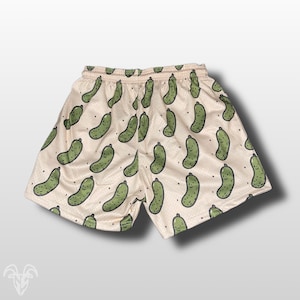 May include: Cream-colored mesh shorts featuring a repeating pattern of smiling green pickle illustrations. The shorts have a drawstring waistband and a relaxed fit, perfect for casual wear or lounging. The pickle design adds a playful and unique touch to the garment.