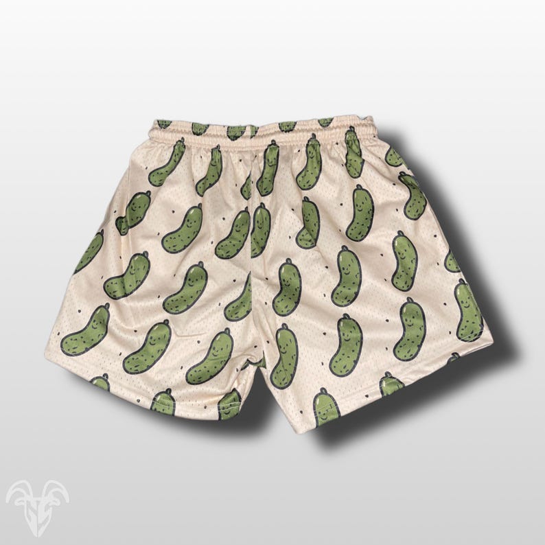 May include: Cream-colored mesh shorts featuring a repeating pattern of smiling green pickle illustrations. The shorts have a drawstring waistband and a relaxed fit, perfect for casual wear or lounging. The pickle design adds a playful and unique touch to the garment.