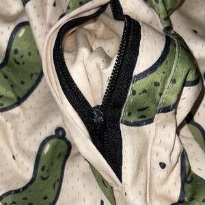 May include: Close-up of a cream-colored fabric with a repeating pattern of green pickle illustrations. A black zipper is partially open, revealing the interior lining. The fabric has a mesh-like texture.