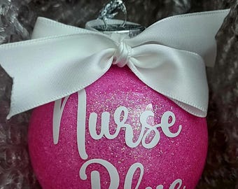May include: A pink glitter Christmas ornament with the words "Nurse Blue" in white script. The ornament has a white satin bow and a silver metal hanger. The ornament is round.