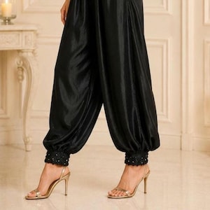 May include: Black satin trousers featuring a loose, voluminous fit with gathered, elasticized cuffs at the ankles. The cuffs are detailed with intricate black lace trim and small, subtle embellishments along the hemline.