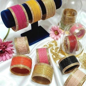 May include: A collection of colorful bangles in various shades, including pink, yellow, gold, orange, and black. Some bangles are displayed on a blue velvet stand, while others are arranged on a white surface. Each bangle is adorned with a decorative border.
