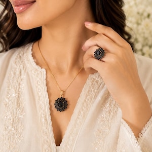 May include: A jewelry set consisting of a gold-tone necklace with a central black oval stone surrounded by small black crystals, matching stud earrings with a similar floral-like design, and a coordinating ring featuring a black oval stone.