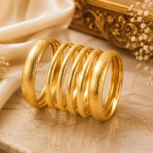 May include: A set of six gold bangles arranged in a row. Each bangle features a textured, matte finish on the exterior surface and a smooth, polished interior. The bracelets are circular in shape and have a consistent thickness throughout the set.