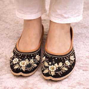 Op de afbeelding: Zwarte geborduurde ballerina's met gouden en parelmoer bloemmotieven. De schoenen hebben een bruine rand en worden gedragen met een witte broek. De ballerina's zijn versierd met kralen en pailletten.