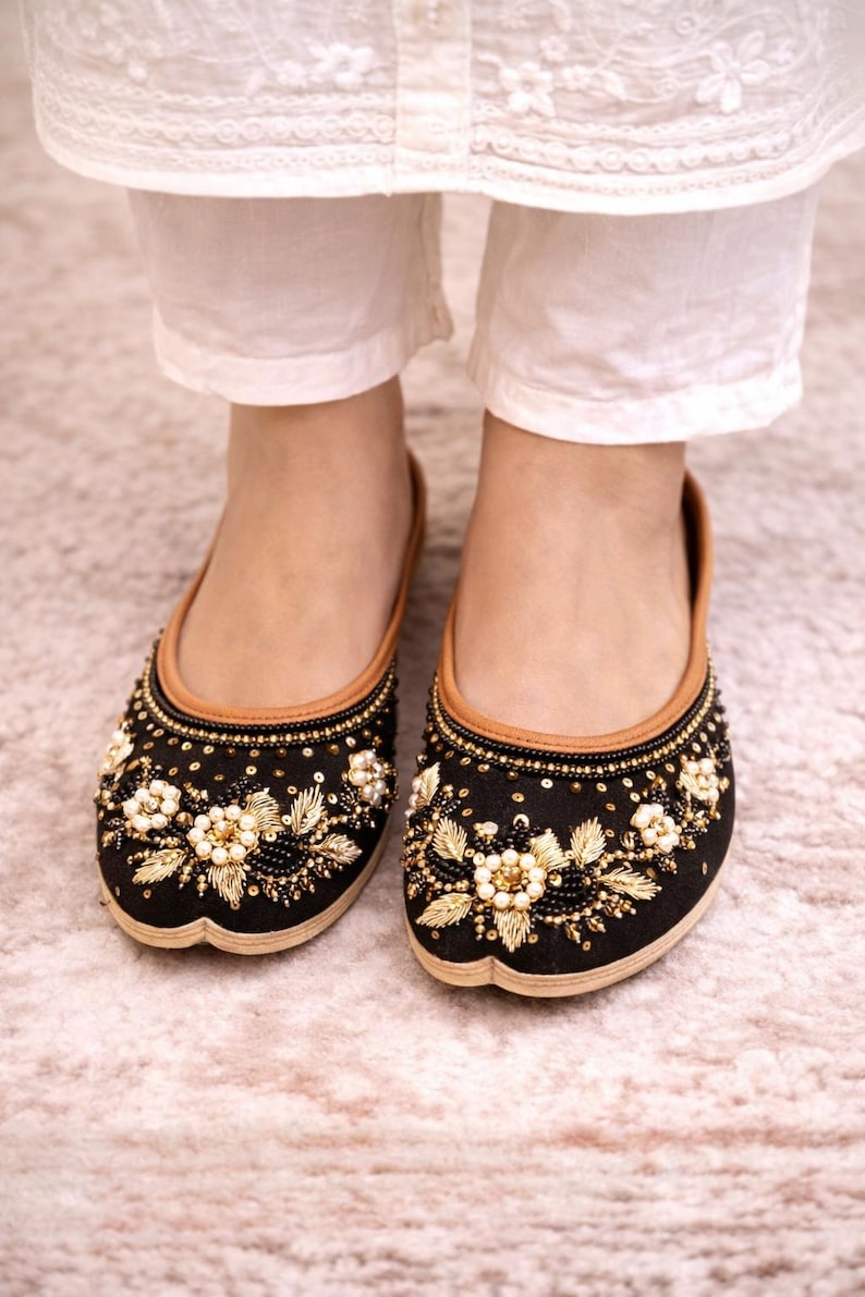 Op de afbeelding: Zwarte geborduurde ballerina's met gouden en parelmoer bloemmotieven. De schoenen hebben een bruine rand en worden gedragen met een witte broek. De ballerina's zijn versierd met kralen en pailletten.
