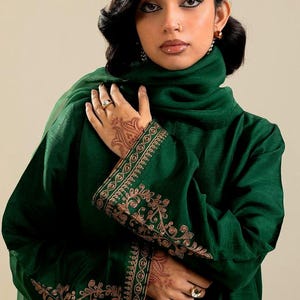 May include: A dark green ensemble featuring a high-neck top and matching scarf. The sleeves are adorned with intricate gold embroidery. The outfit is worn by a person with henna art on their hand and a ring.