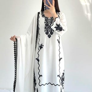 May include: A white long-sleeved tunic featuring intricate black floral embroidery around the neckline, cuffs, and hem. The garment is paired with a matching white dupatta that has a decorative black lace border along the edges and white trousers.