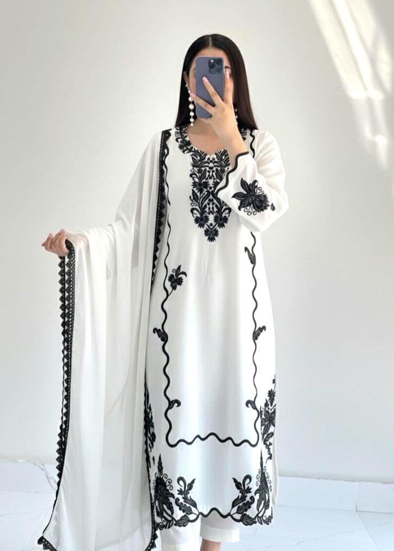 May include: A white long-sleeved tunic featuring intricate black floral embroidery around the neckline, cuffs, and hem. The garment is paired with a matching white dupatta that has a decorative black lace border along the edges and white trousers.