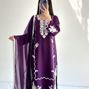 May include: A deep purple long-sleeved tunic featuring intricate white floral embroidery around the neckline, cuffs, and hemline. The outfit includes matching purple trousers and a sheer purple dupatta scarf with a decorative white scalloped lace border.