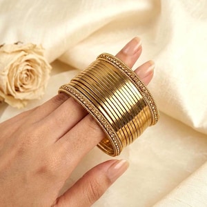 May include: A stack of gold-colored metal bangles featuring a smooth, polished finish on the central bands and a textured, beaded pattern on the two outer bangles. The set consists of multiple thin, circular bracelets worn together as one unit.