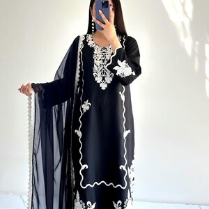 May include: A black long-sleeved tunic featuring intricate white floral embroidery around the neckline, cuffs, and hem. The garment is paired with a matching black sheer dupatta that has a decorative white scalloped lace border along the edge.
