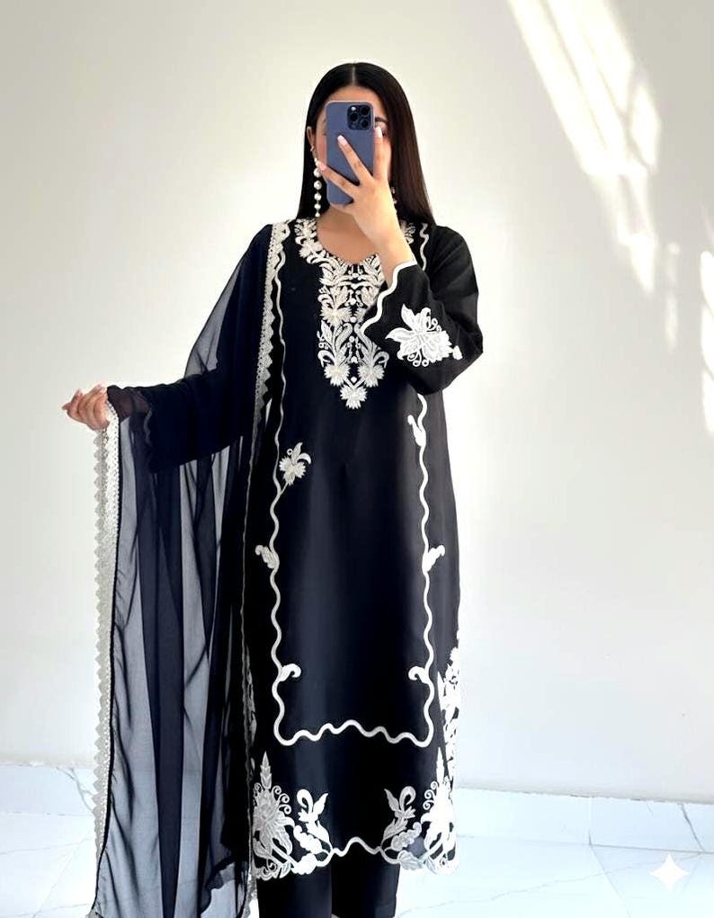 May include: A black long-sleeved tunic featuring intricate white floral embroidery around the neckline, cuffs, and hem. The garment is paired with a matching black sheer dupatta that has a decorative white scalloped lace border along the edge.