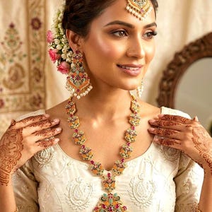 May include: An ornate gold necklace, earrings, and headpiece set with colorful gemstones and pearls. The jewelry is worn with a cream-colored embroidered top. The woman has henna designs on her hands and wrists.