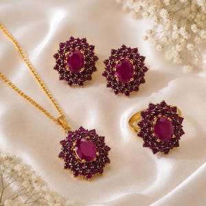 May include: A jewelry set consisting of a gold necklace with a pendant, a pair of stud earrings, and a ring. Each piece features a large oval-cut deep pink gemstone surrounded by a halo of smaller round-cut pink stones in a gold setting.