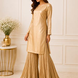 May include: A metallic gold long-sleeved tunic top paired with matching wide-leg pleated palazzo pants. The tunic features a round neckline and side slits, while the floor-length pants have a dense, vertical accordion-pleated texture throughout.