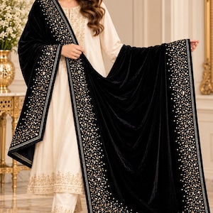 May include: A cream-colored long-sleeved tunic and matching trousers featuring gold embroidery at the neckline and hem, paired with a black velvet shawl adorned with gold sequins, beads, and metallic accents along the wide borders.
