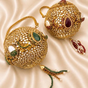 May include: Two spherical gold-toned handbags featuring intricate metallic filigree, embedded with numerous clear crystals, and accented with large teardrop-shaped gemstones in deep green and dark red, finished with beaded tassels and handles.