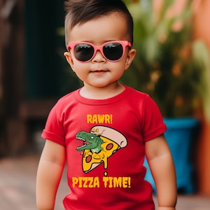 May include: A red toddler t-shirt featuring a graphic of a dinosaur eating a slice of pizza. The text on the shirt reads "RAWR! PIZZA TIME!"