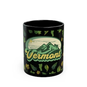 May include: Black ceramic mug with a green and yellow design featuring a mountain range and the word "Vermont". The background is black with green leaves.