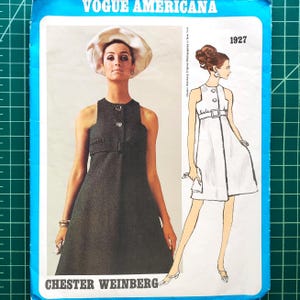 Op de afbeelding: Een vintage Vogue Americana naaipatroon. De afbeelding toont een vrouw in een donkergrijze jurk met een riem en een witte hoed. Er is ook een lijntekening van een witte jurk opgenomen. De tekst "CHESTER WEINBERG" staat onderaan.
