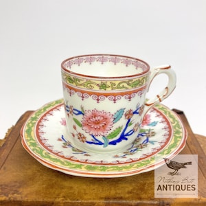 Puede incluir: Un juego de taza y platillo vintage. La taza y el platillo son blancos con un diseño floral en rosa, azul y verde. Los bordes están adornados con detalles rojos y amarillos. El asa de la taza es curvada.