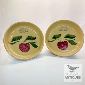 Puede incluir: Dos platos antiguos de cerámica amarilla con diseño de cereza. Cada plato presenta una cereza roja con hojas verdes y el texto "Farmers Union Co-Op Hooper, Nebr." en negro. Los platos tienen un borde ligeramente elevado.