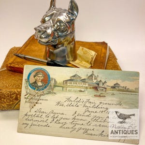 Peut inclure: Un encrier en métal argenté en forme de tête de chien repose sur un livre ancien à couverture en cuir brun, accompagné d'un stylo plume noir. Au premier plan, une carte postale vintage de l'Exposition universelle de 1893 illustre un bâtiment.