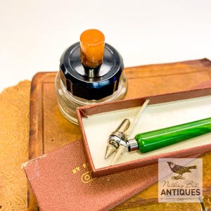 May include: An antique inkwell with a black lid and orange stopper sits next to a vintage green pen in a brown box. The pen has a silver nib. The items rest on a stack of old books. The image includes the text "Nothing But Antiques".