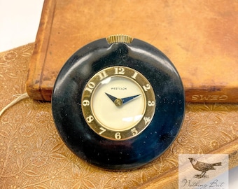 Könnte beinhalten: Eine runde Westclox-Reiseuhr mit schwarzem, kreisförmigem Gehäuse und goldfarbener Lünette. Das Zifferblatt ist weiß mit schwarzen Ziffern von 1 bis 12 und zwei dunklen Zeigern. Ein kleiner goldfarbener Knopf befindet sich oben am schwarzen Gehäuse.