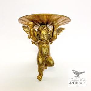 May include: A golden cherub wall shelf. The cherub figure is holding up a large, shell-shaped shelf. The cherub has wings and is kneeling. The shelf is a decorative piece for displaying small items. The finish is a metallic gold.