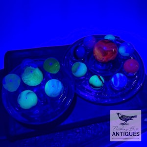 May include: A collection of colorful glass marbles in various sizes, displayed in clear glass dishes. The marbles feature swirls of red, blue, green, and yellow. The image is set against a dark blue background, with the text "Nothing But ANTIQUES".