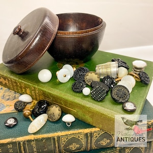 May include: A wooden box with a lid, open to reveal a collection of black, white, and gold buttons. The box is sitting on top of two antique books. The books have a green cover and gold lettering. The image includes the text "Nothing But Antiques".