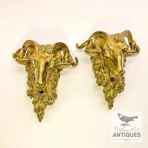 May include: Two antique brass ram head wall sconces. The ram heads have curled horns and textured fur, and are mounted on a white background. The sconces are a golden colour and appear to be vintage. The image includes the text "Nothing But Antiques".