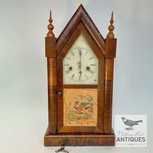 Puede incluir: Un reloj de madera con forma de campanario y acabado marrón oscuro, que presenta una esfera blanca con números romanos negros y dos manecillas de metal. Debajo de la esfera, un panel rectangular muestra una ilustración colorida de un ave.