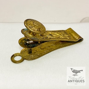 Puede incluir: Hebilla de cinturón antigua de latón con detalles florales y de volutas ornamentales. La hebilla presenta un botón circular con texto en relieve y un diseño articulado. El metal tiene un aspecto deslustrado y envejecido, lo que sugiere su origen vintage. La hebilla es probablemente de principios del siglo XX.