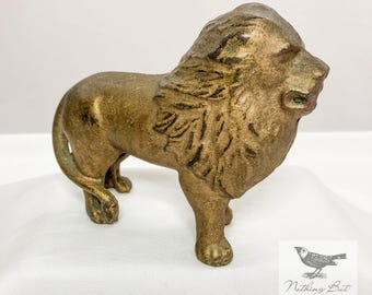 May include: A bronze-coloured lion figurine with a detailed mane and a regal pose. The antique sculpture has a textured surface and is standing on a white surface. The lion's features are well-defined, showcasing its powerful form.