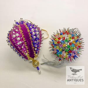Puede incluir: Dos adornos navideños decorativos. Uno es una forma de lágrima de terciopelo morado oscuro, adornada con cuentas y perlas. El otro es una esfera plateada cubierta de cuentas de colores. Ambos tienen lazos para colgar.