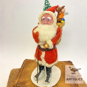May include: A vintage Santa Claus figurine wearing a red felt coat with white faux fur trim, a matching hat, and black boots. Santa carries a woven basket on the back containing a small wooden angel, a brown deer, and a green bottle brush tree.
