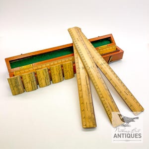 May include: Antique wooden slide rule set with a wooden box. The set includes multiple wooden rulers and a box with a green felt interior. The rulers are marked with measurements in inches and other markings. The box is brown with a brass clasp.