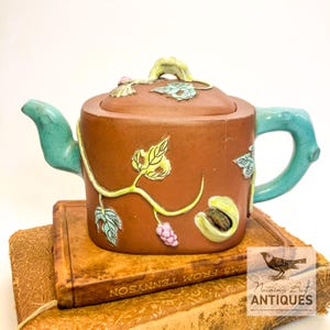 Può includere: Una teiera in ceramica marrone con coperchio, manico e beccuccio turchese e dettagli di vite e foglie. La teiera è decorata con accenti gialli, verdi e rosa. La teiera è appoggiata sopra un libro d'epoca.
