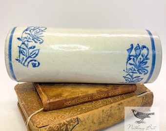 May include: A cylindrical ceramic rolling pin with a cream-colored glaze and blue floral patterns on each end. The rolling pin is positioned horizontally on top of two stacked antique books, one of which is titled Gems from Tennyson.