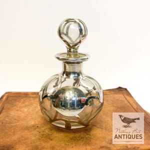 May include: A vintage perfume bottle with a silver-colored stopper and accents. The clear glass bottle has a round shape with a silver emblem. The bottle is sitting on a brown surface. The logo "Nothing But Antiques" is in the bottom right corner.