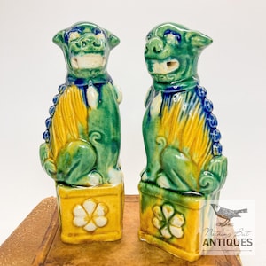 May include: Pair of ceramic Foo Dog statues. Each statue is green, yellow, and blue, with a detailed design. The Foo Dogs are seated on a yellow base with a white flower design. The statues are displayed on a brown surface.